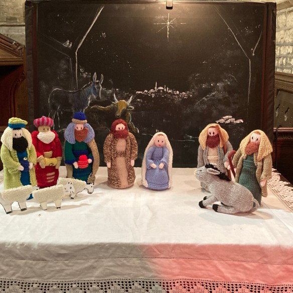 Woolly Nativity of St Peter's Church, shows the knitted figures of the Christmas story against a painted backdrop with the animals in the stable and views beyond to Bethlehem and the Star of the East