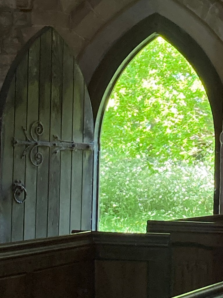 View through the North Door - St Peter's Church. Image shows dark interior of old church, with large wooden door open to the left. Outside is the view to the bright greens of the trees in the churchyard. A beautiful bright view