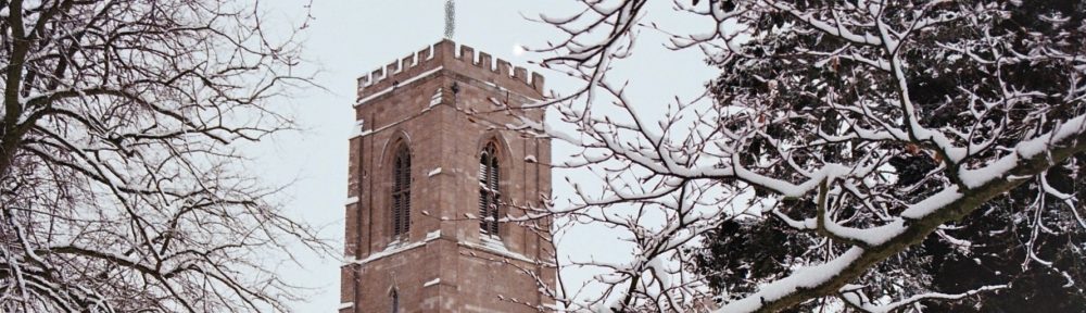 Photo of St Peter's church in the snow - the stone work looks reddish pink agains the white snow and the bare trees frame the building and tower