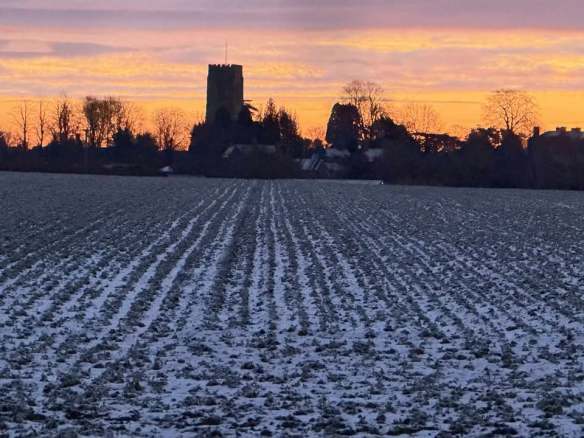 Winter view of St Peter's Church Langton. The building is silhouetted against the setting sun, with a snow lined ploughed field in the foreground. The furrows lead to the church in a single point perspective
