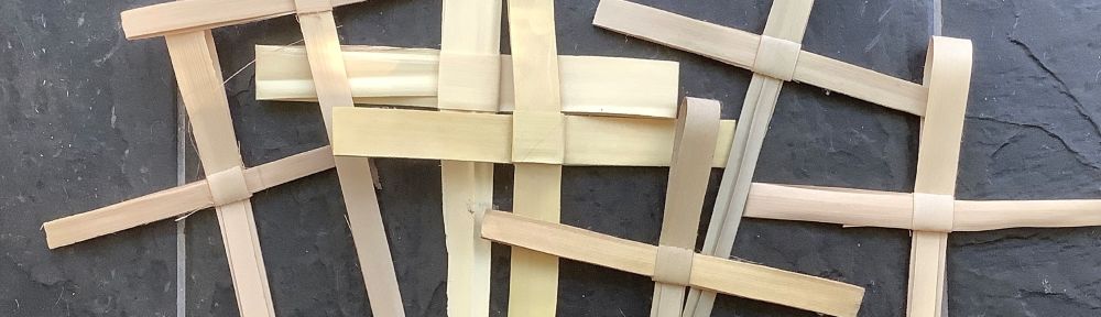 Photo by Maxine Dodd of a collection of seven Palm Sunday crosses, against a black background. The crosses are handmade from fronds of real palm trees, used by the people to greet Jesus as he arrived in Jerusalem at the start of Holy Week.